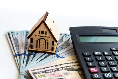 wooden model of house on stack of cash next to calculator